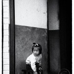 A small Chinese girl sits obediently in a hallway as she keeps an anxious eye out for her mother.
